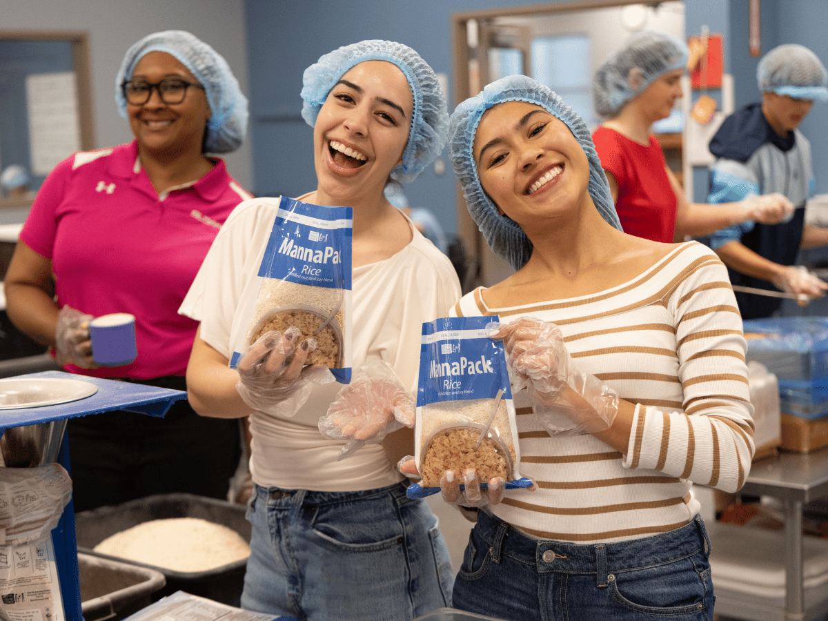 Two girls in hairnets hold up a MannaPack meal they packed at an FMSC volunteer packing session